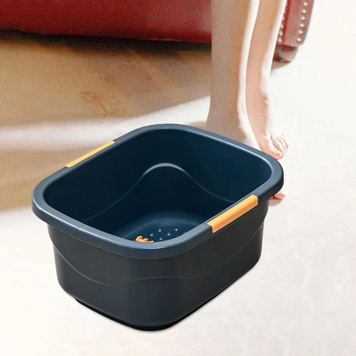 Bain de pied - Bassin portable de couleur noire avec poignées orange, vue en perspective sur un sol clair, pieds nus à proximité.