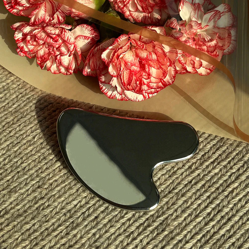Appareil massage visage en acier inoxydable – forme ondulée avec finition brillante, posé sur un tissu beige à côté de fleurs roses