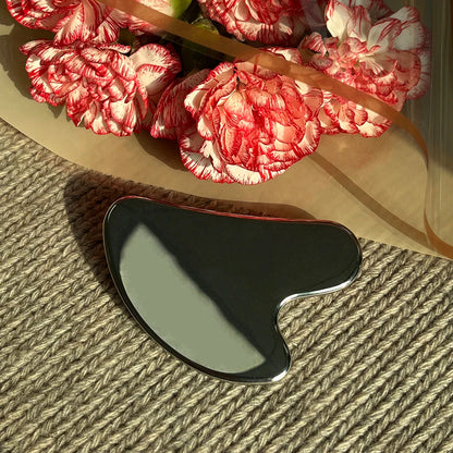 Appareil massage visage en acier inoxydable – forme ondulée avec finition brillante, posé sur un tissu beige à côté de fleurs roses