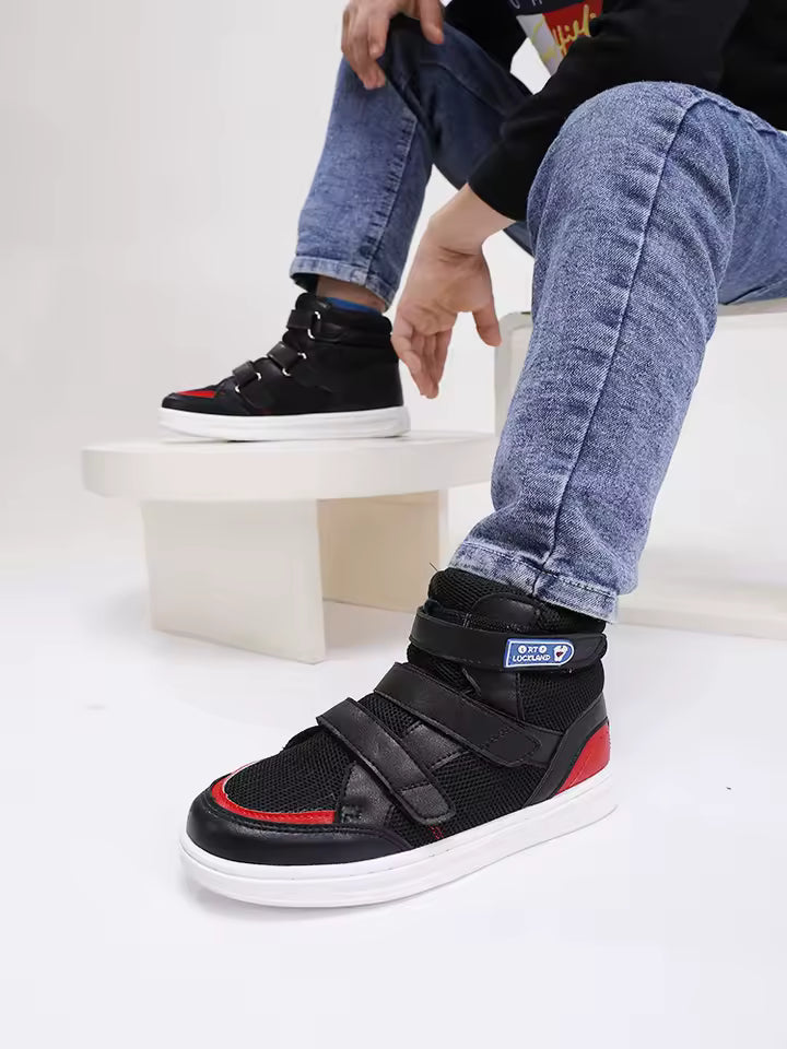 Chaussures orthopédiques enfant FlexiFit noires avec détails rouges, portées par un enfant assis, vue latérale mettant en avant les bandes velcro.