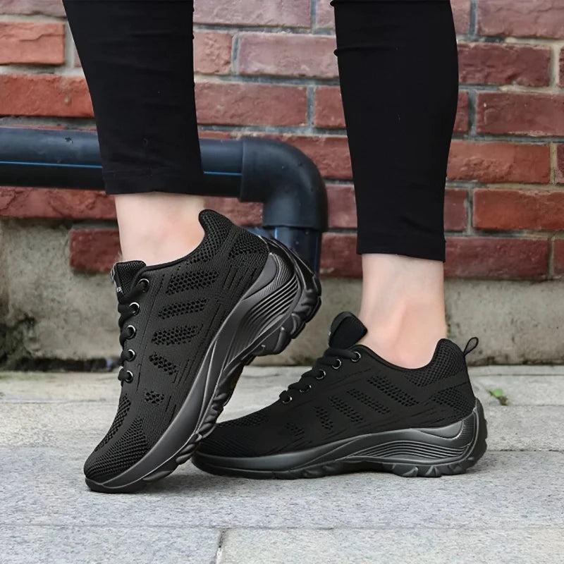 Basket orthopédique femme - Chaussures de sport respirantes en maille noire avec détails en relief, portées sur un sol en pavés.