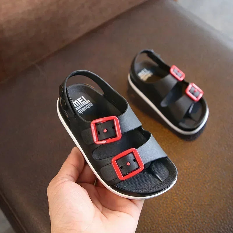 Chaussure de plage enfant noire avec deux boucles rouges, vue de près tenue dans une main, sur une surface marron.