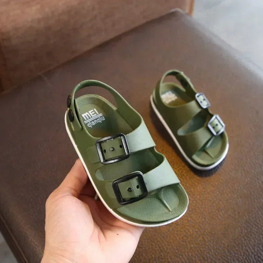 Chaussure de plage enfant verte avec deux boucles, vue de près dans une main, sur une surface marron.